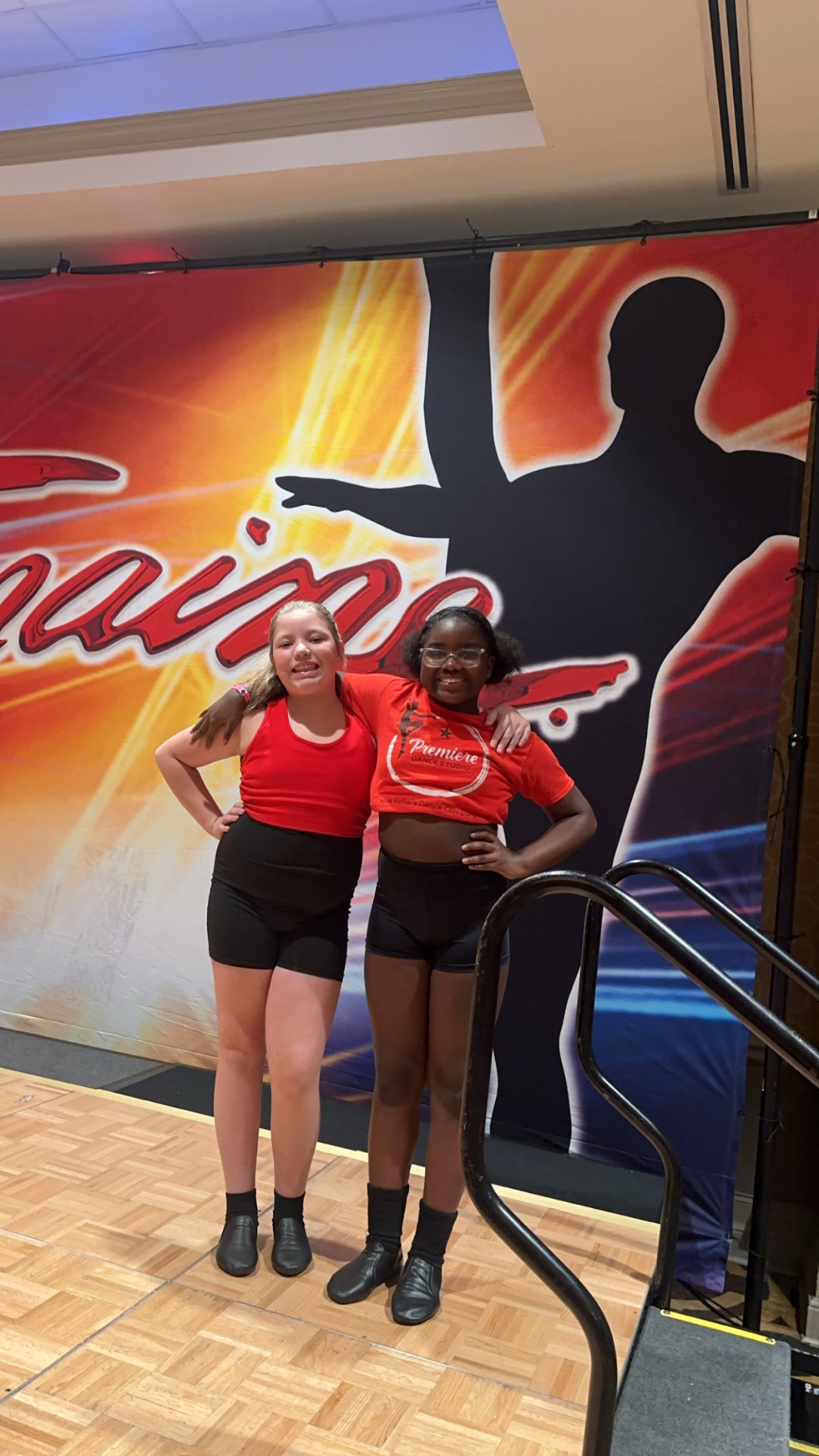 Two Premiere dancers in red tanks at a Tremaine competition backdrop.