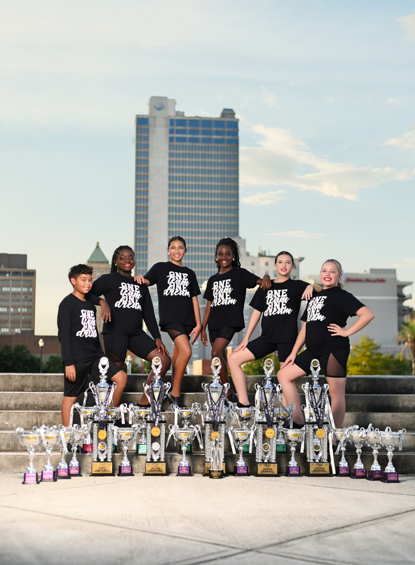 Premiere dancers in One Team One Dream shirts posing with a wall of trophies